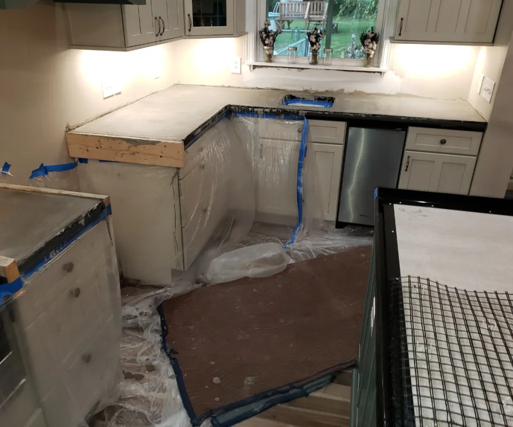 Kitchen counters mid-install over new cabinets, the floor covered and the sink plumbing roughed in